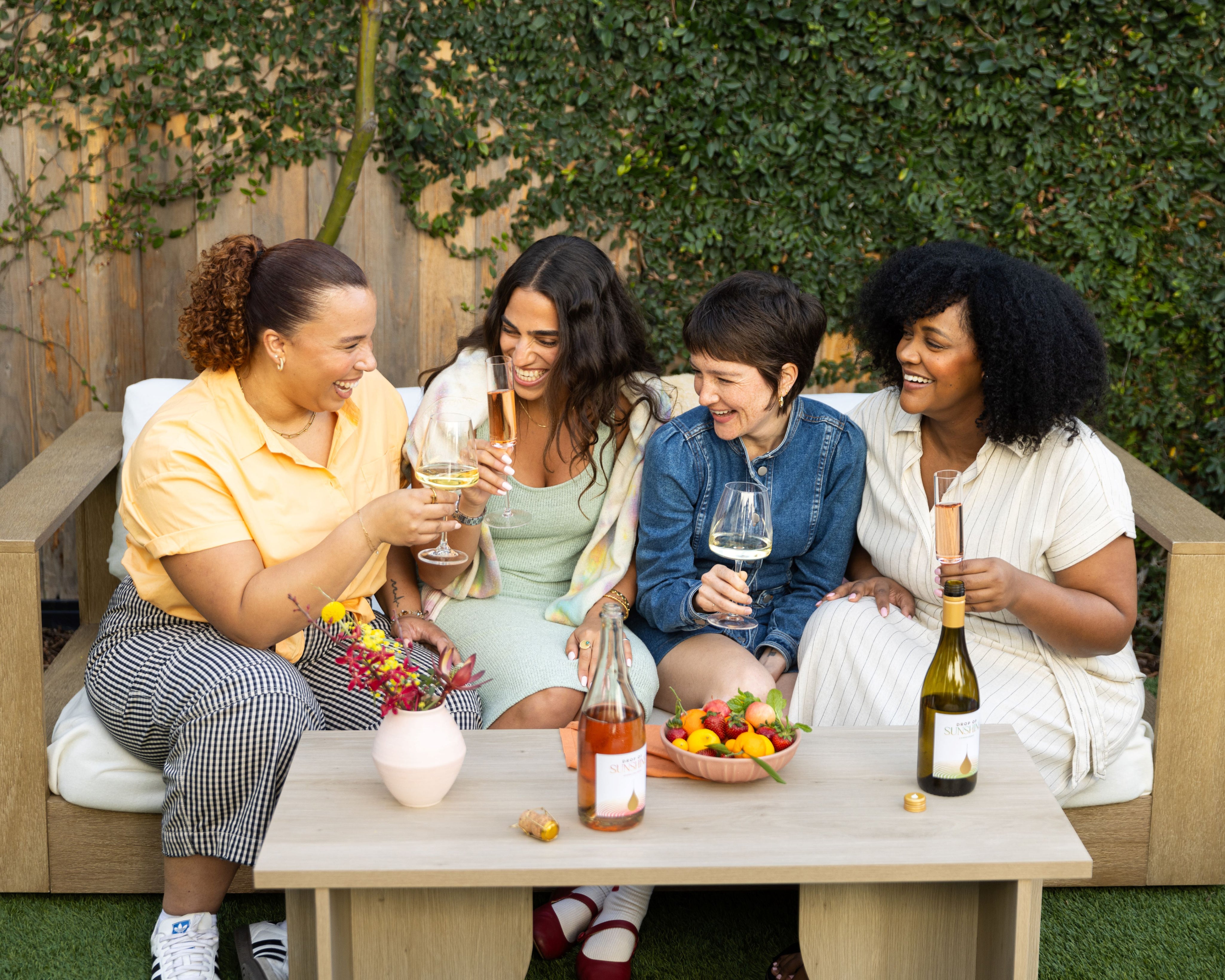 Four women sitting on a bench each with a glass of wine celebrating by bringing their glasses together in a manner post cheers