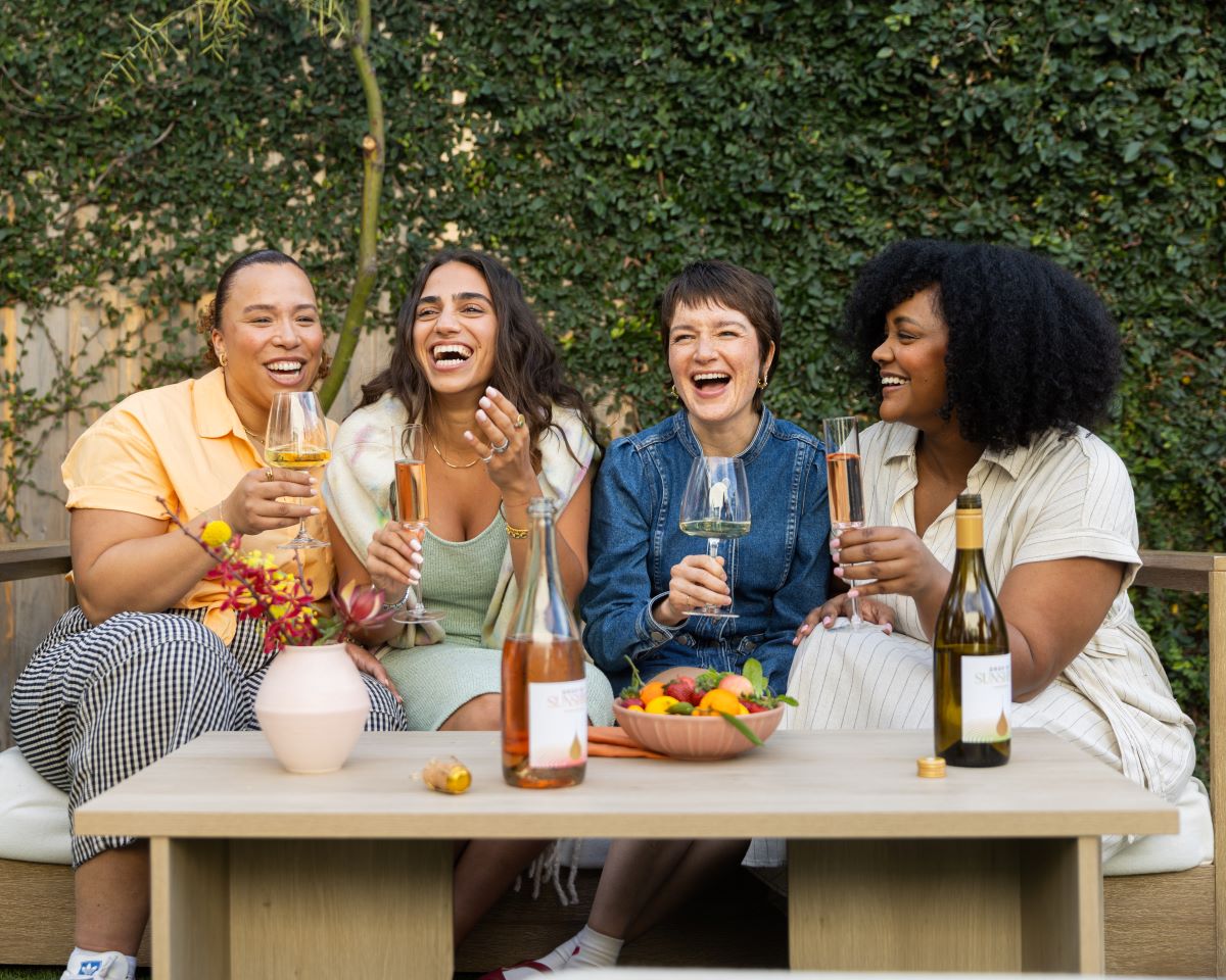 Four women sitting on a bench each with a glass of wine celebrating by bringing their glasses together in a manner post cheers and laughing