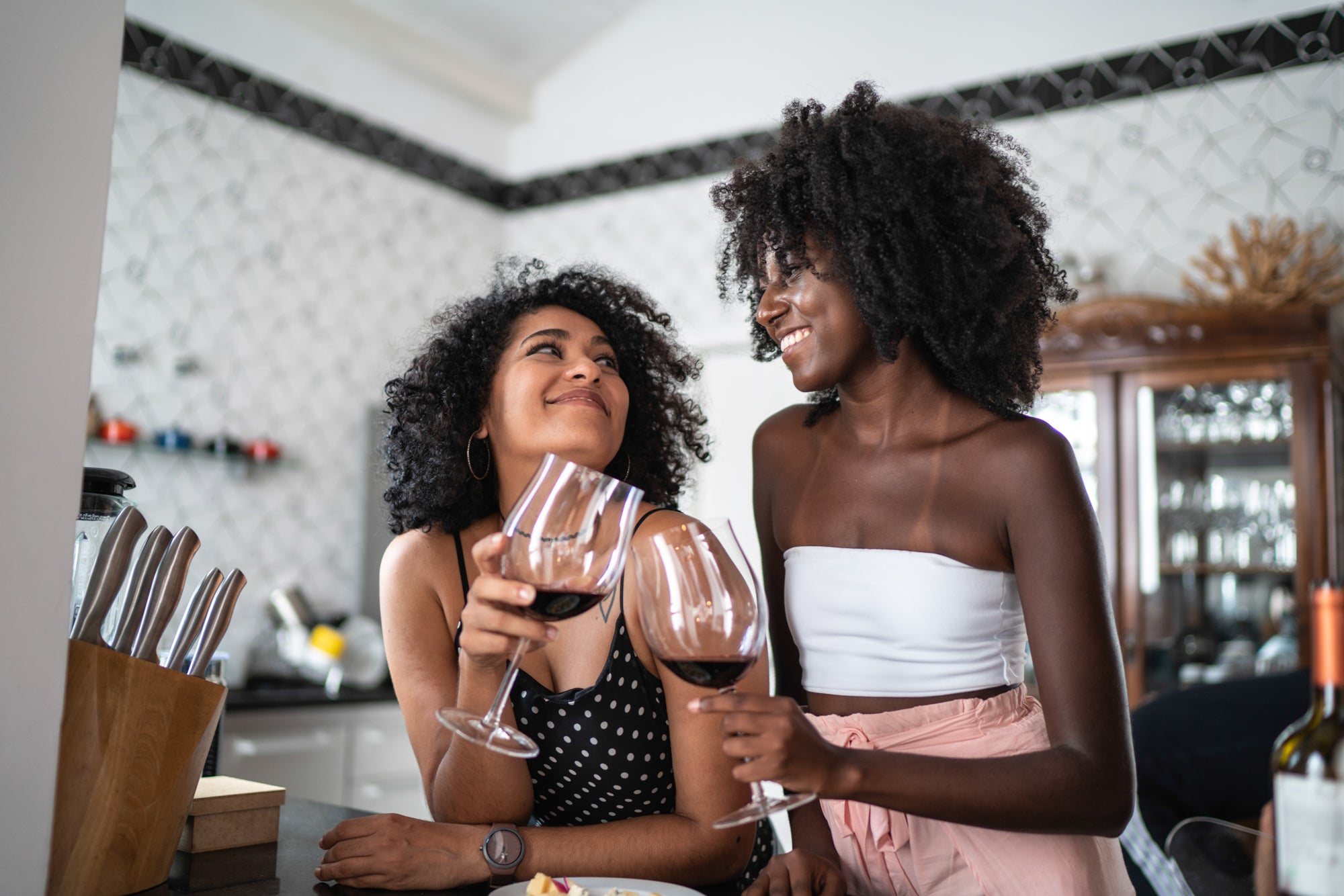 Two women smile at each other and cheers red wine in a beautiful kitchen